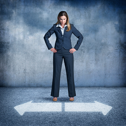 A businesswoman with hands on hips standing and slightly bent over, looks down at two arrows pointing in opposite directions. She is confronted with having to make the choices that are common in everyday life.
