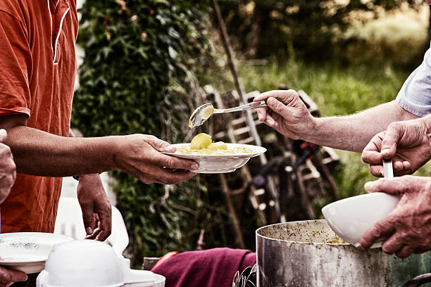 soup serving kitchen soup kitchen serving food for poor people. One man is holding his plate out for a serving. The person serving is holding a spoon and serves soup and potatoes. The man receiving the food is wearing a reddish dress with short sleeves. soup kitchen stock pictures, royalty-free photos & images