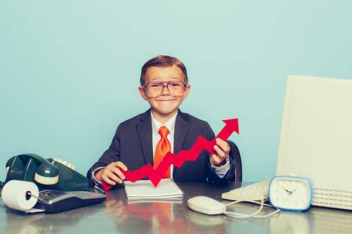 A young boy dressed as a businessman sits at his desk smiling as there is some good business happening. He sits at his desk with retro computer and phone.