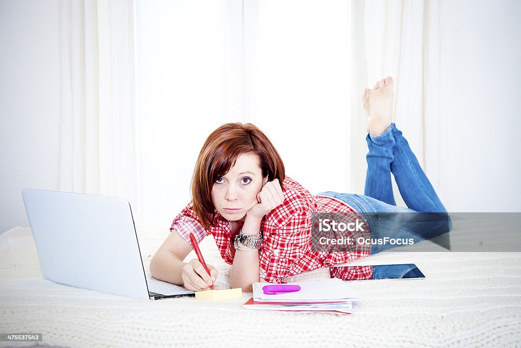 bored red hair student, business woman in bed with computer bored attractive red hair student or business woman lying down on bed working on her computer wearing a casual red shirt and jeans on white background Adult Stock Photo