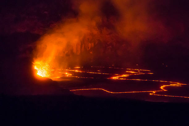 Orange molten magma erupts at night inside Hawaii Island's Kilauea volcano. The Lava lake is glowing orange inside a volcanic caldera. A cloud of volcanic gasses is illuminated by glowing molten lava splattering into the air. The night image is horizontal and mostly dark except for the glowing areas.