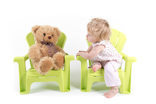 Baby Girl sitting on green garden chair and taking with toy bear on other chair, studio isolated, white background