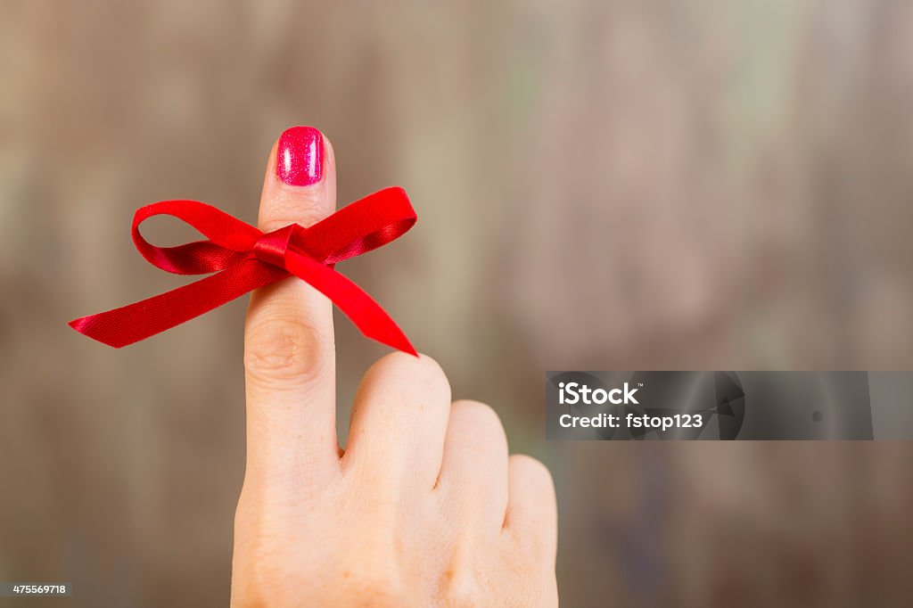 Red social awareness ribbon tied around index finger. Reminder. Red social awareness ribbon tied in a bow around a woman's index finger.  The red ribbon represents various causes including: AIDS/HIV, heart disease, stroke, and substance abuse. Gray background. Copyspace. Ribbon - Sewing Item Stock Photo