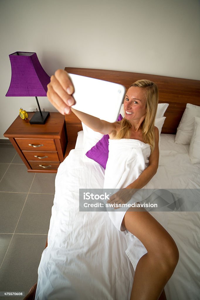 Young woman taking a selfie on bed-Bedroom Young woman on a comfortable bed in an hotel room taking a selfie using her mobile phone. Sunlight coming in the room. Bed - Furniture Stock Photo