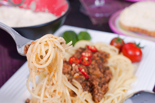 Cooked spaghetti with ground beef meat, pasta sauce, red chili peppers and cherry tomatoes, served with a fork on a white rectangular plate, close-up