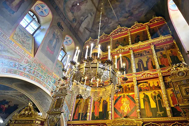 Interior of Russian Orthodox Church. A view of an iconostasis, walls and a ceiling with frescos and a suspended chandelier.