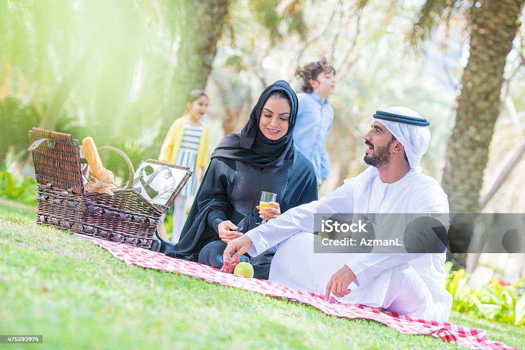 Picnicing in a Park Arab family having picnic in a park. Parents are wearing traditional emirati clothes. They are sitting on a blanket, talking, drinking juice, kids are playing in back. 2015 Stock Photo