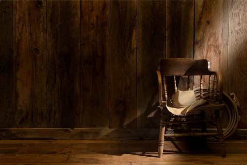 This image has subdued rustic barnwood with a vintage saloon chair, cowboy hat and lasso. A shaft of soft sun ray comes in from the right to highlight the chair and hat. Room for overprint