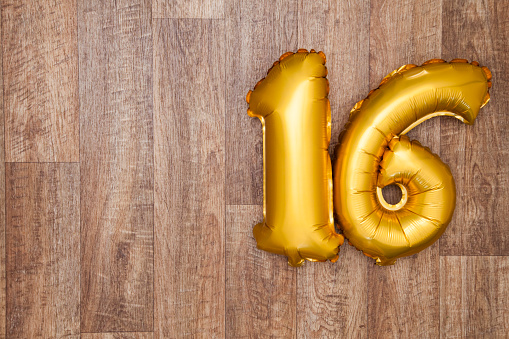 A gold foil number 16 balloon on a wooden background. The number is made from shiny golden foil and is inflated, it is on the right hand side of the image leaving copy space on the left of the image for your text or logo.