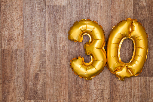 A gold foil number 30 balloon on a wooden background. The number is made from shiny golden foil and is inflated, it is on the right hand side of the image leaving copy space on the left of the image for your text or logo.