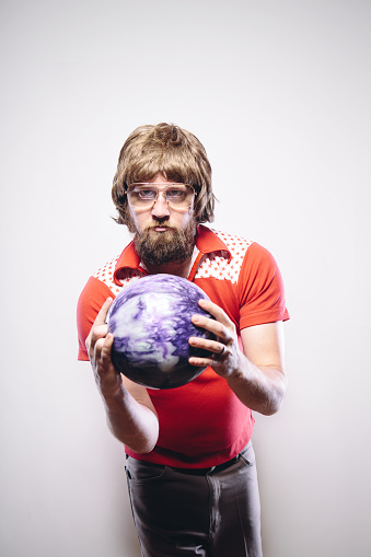 A shaggy haired man with a beard, glasses, and classic retro bowling shirt holds his ball, ready to bowl.  Vertical portrait with copy space above him.  Off white background with vignette.
