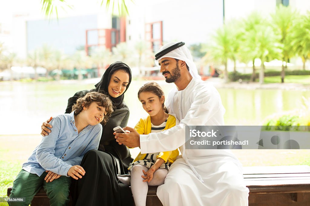 Arab family in the park Traditional arab family taking selfie in the park. The man is   worn in traditional male dress kandura (dishdasha) with kaffiyeh (white headdress of cotton fabric) held in place by a rope (agal).He is holding mobile phone. The woman is worn  an Abaya and Hijab, the traditional national dress of the United Arab Emirates, Saudi Arabia, Kuwait, Bahrain, Iraq, Oman, Qatar and Yemen. Image is taken during Dubai istockalypse. Eid-Ul-Fitr Stock Photo