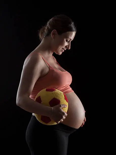 Pregnant Woman Holding Soccer Ball In Red And Yellow Color.Model is looking toward right side of frame and touching her belly.Background is black.Main light is on the right side of frame and outline border is enlightened with a back lit.The photo was shot with a full frame DSLR camera in studio.