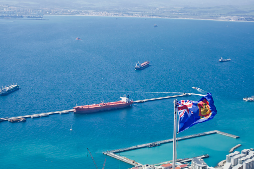 Cargo ships around Gibraltar harbor, Gibraltarian flag in the foreground and spanish coastline in the background.