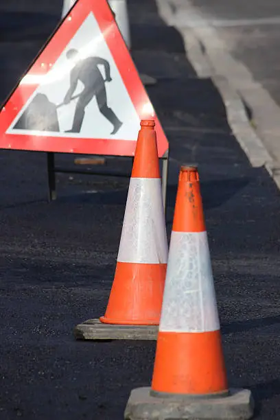 Photo showing a pavement that has just been resurfaced with tarmac.  As the layer of asphalt is still hot, the sidewalk features a series of bollards and a triangular 'men at work' traffic road sign.