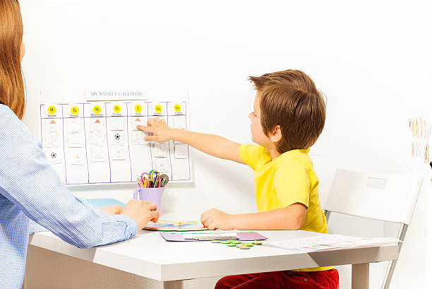 Boy points at activities on calendar learning days Boy pointing at the calendar on the wall with days and activities arranged developing game with his parent sitting opposite at the table indoors personal organizer photos stock pictures, royalty-free photos & images