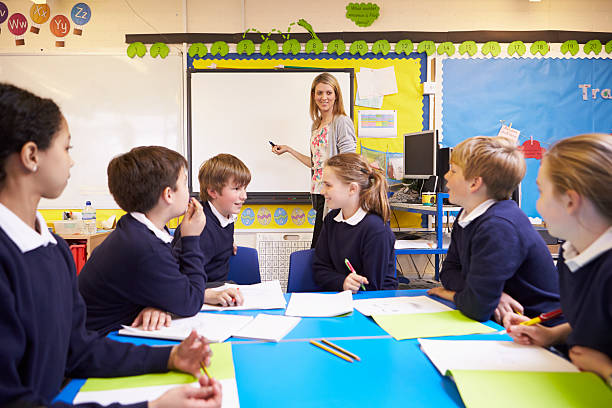 istock Pupils Sitting At Table As Teacher Stands By Whiteboard 475684133