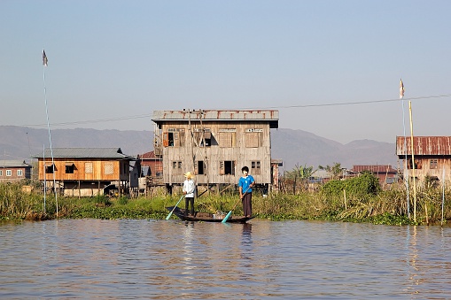 Nyaung Shwe township, Myanmar - February 5, 2015: Burmese men are going by traditional boat on the Inle Lake, with the village overwater houses in the background