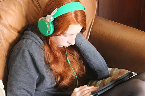 Photo showing a young girl with red hair, sitting on an old brown leather sofa and wearing green headphones, as she listens to music and watches videos on her tablet computer device in the lounge.