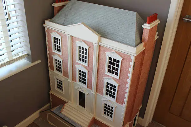 Photo showing a beautiful homemade toy wooden Georgian dollshouse, which features a part red-brick, part painted cream exterior.  Many grand, elegant elements make up the facade, including panelled windows, a front door with a portico, chimney pots and steps.
