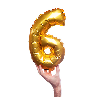 A gold foil number 6 balloon is held by a caucasian male hand. The number is being held at the base. The number is made from shiny golden foil and is inflated. The background is plain white.