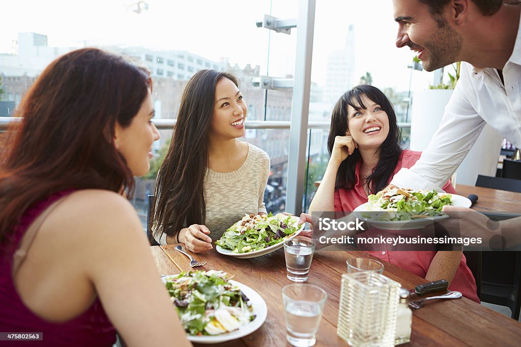 Three Female Friends Enjoying Lunch At Rooftop Restaurant Three Female Friends Enjoying Lunch At Rooftop Restaurant Smiling And Laughing With Each Other. Waiter Stock Photo