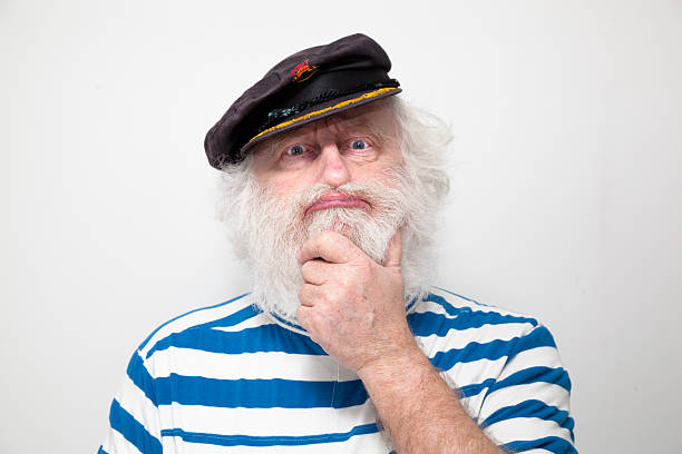 bearded senior sailor with cap and striped jersey  puzzled and scratching beard - on plain background