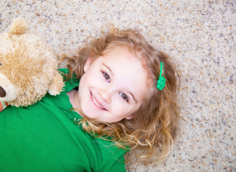 Young girl laying on the floor with a teddy bear, smiling up at the camera.