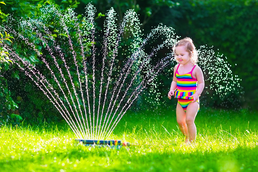 Child playing with garden sprinkler. Kid in bathing suit running and jumping. Kids gardening. Summer outdoor water fun. Children play with gardening hose watering flowers.
