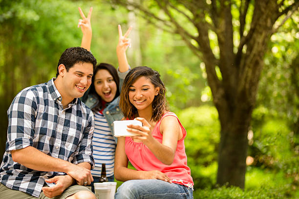 Young adult friends take 'selfie' using smart phone. Photo bomb. Two young adult friends take a 'selfie' photo using their cell phone. The woman is holding the smart phone while the other friend smiles for the camera.  A woman in the background is "photo bombing the photo."  Latin descent, caucasian, and mixed-race group of people.  Beautiful spring or summer nature background.  Park, campus, or backyard setting. photo bomb stock pictures, royalty-free photos & images