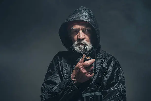 Portrait of a Reflective Bearded Old Guy in Black Rain Jacket, Smoking a Cigarette Using a Pipe. Captured in Studio.