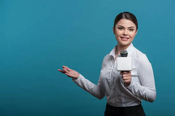 Waist up portrait of elegant woman reporter with brown hair, who interviews and is smiling and looking at the camera holding the microphone and making a gesture with her right hand, isolated on a blue background and there is copy place in the left side