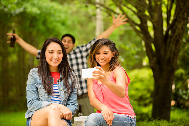 Young adult friends take 'selfie' using smart phone. Photo bomb. Two young adult girl friends take a 'selfie' photo using their cell phone. The woman to right is holding the smart phone while the other friend smiles for the camera.  A man in the background is "photo bombing the photo."  Latin descent, caucasian, and mixed-race group of people.  Beautiful spring or summer nature background.  Park, campus, or backyard setting. photo bomb stock pictures, royalty-free photos & images