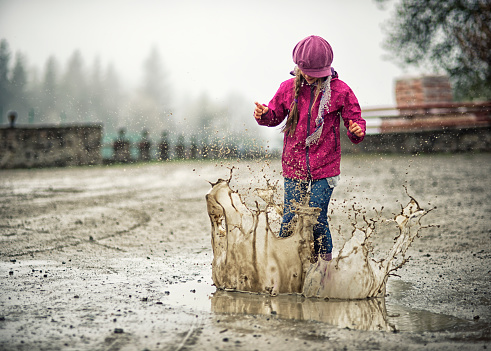 Little girl in galoshes jumping into a spring puddle. There is a big splash of dirty water.