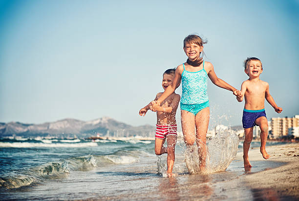 Happy kids running on a beach. The girl aged 7 is holding hands of her two little brothers aged 4. Three kids are running on a beautiful beach splashing and laughing. Mallorca, Spain.
