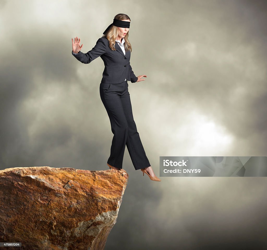 Blindfolded businesswoman about to step off of a clif A blindfolded businesswoman about to step off of a ledge. She is surrounded by stormy skies.  Room for text. Cliff Stock Photo