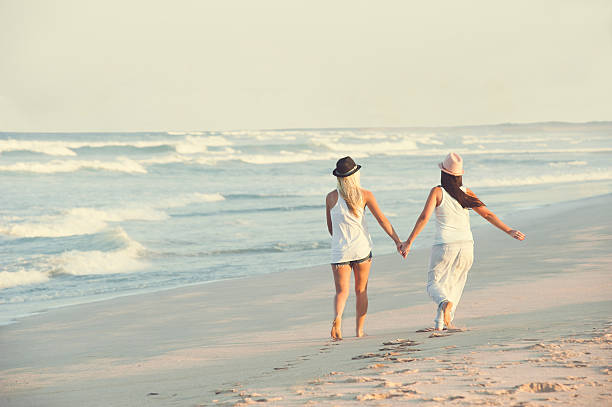two young girls holding hands walking along beach stock photo