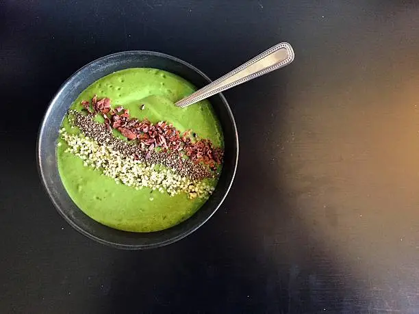 A top-down view of a green smoothie bowl topped with chia seeds, cacao nibs and hemp seeds, served in a black bowl with a spoon. Smoothie bowls are a trendy alternative to smoothies served in a glass. They are thicker and eaten with a spoon. This one contains banana, mango, pineapple, kale and hemp seeds.