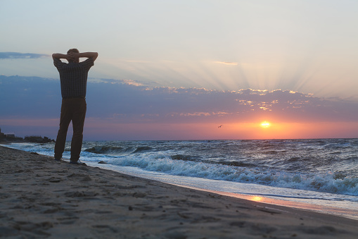 Man stands on the beach and watching sun come up through the clouds on the sea horizon