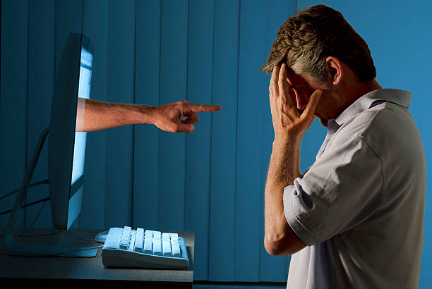 Cyber internet computer bullying and social media stalking Severely distraught young man sitting in front of a computer with a judgmental hand pointing at him from within the computer monitor which shows the man being either computer bullying bullied or Facebook social media stalking stalked. adulation stock pictures, royalty-free photos & images