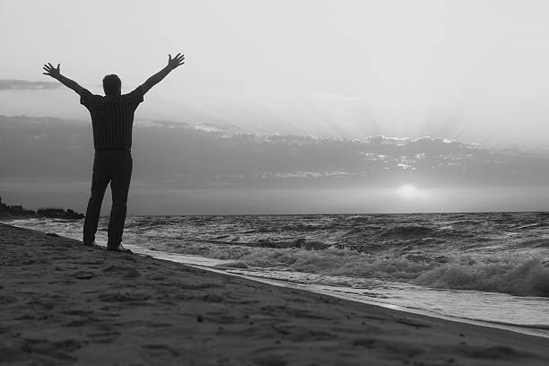 Monochrome image. Man raised his hands gleefully toward the rising sun on sea beach