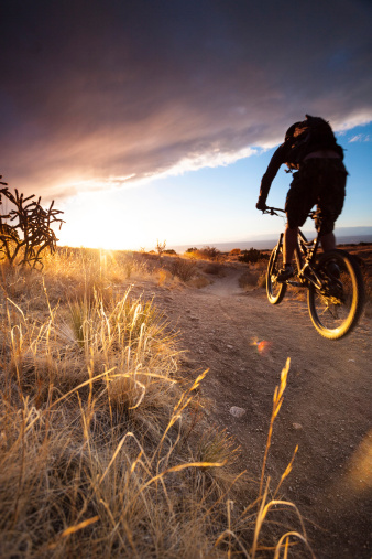 a mountain biking man jumps while riding fast through a desert sunset landscape with dramatic sky and sunlight. such beautiful nature scenery and outdoor sports, fitness, and recreation can be found in the sandia mountains of albuquerque, new mexico. horizontal wide angle composition.