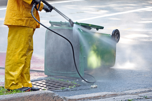 Sanitation worker uses a power washer to clean a compost recycling green bin.