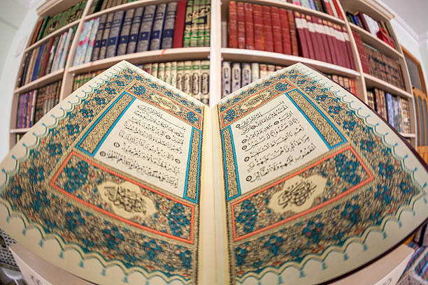 Wide Angle View Of Holy Quran In Library.The Quran is placed in front of book shelf in library.First page of Quran with ornaments around is open.No people is seen in frame.Focus is on Quran.The photo was shot with a full frame DSLR camera and a fisheye lens.