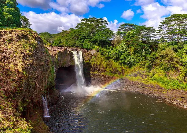Waterfall with rainbow in Hawaii, also know as Waianuenue Falls that flows 80 feet over a natural lava cave into a turquoise pool of water surrounded by dense forest.