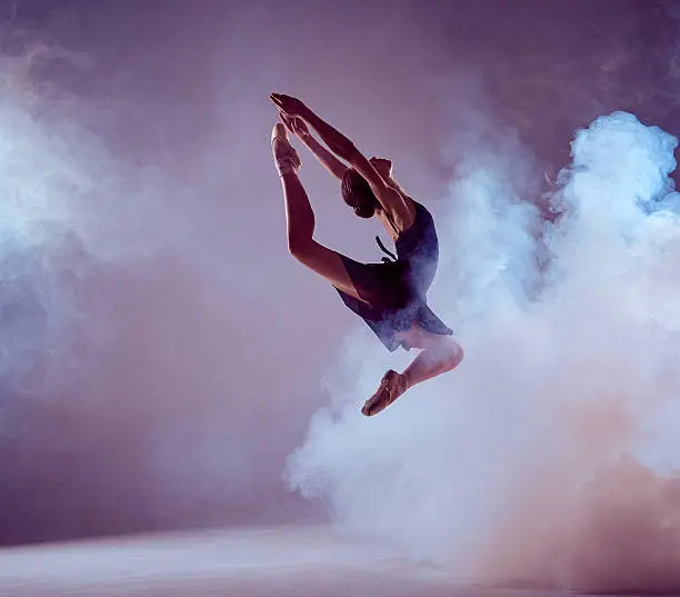young ballet dancer jumping on a lilac background. Ballerina is wearing in blue dress and pointe shoes. The outline shooting - silhouette of girl with smoke effect