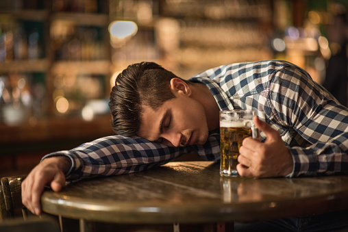 Young exhausted drunk man holding a glass of beer and sleeping on the table in the pub.