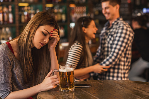 Young depressed woman sitting in the bar and holding her head in pain while happy couple is laughing in the background.