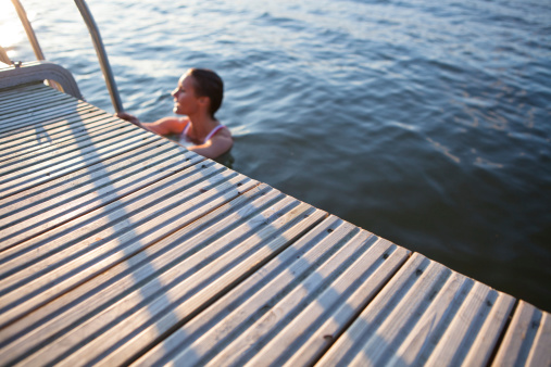 Edge of wooden pier in focus with woman in water holding on to ladder