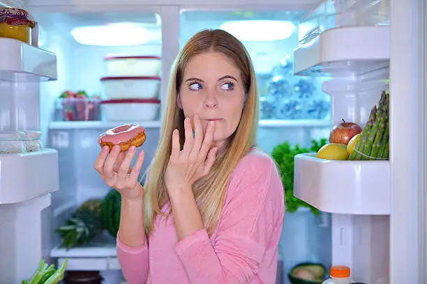 Young woman standing in the refrigerator door and eating donut at night while looking around if someone sees her.